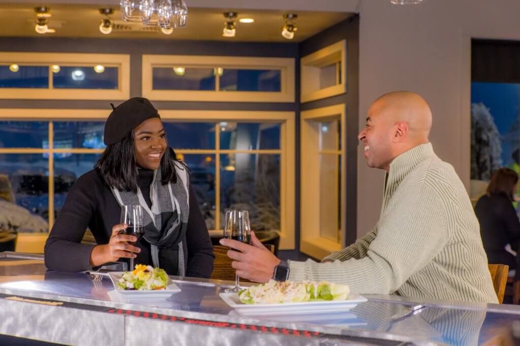 A man and a woman sitting at a bar holding glasses of wine with plates of food in front of them